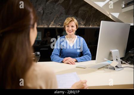 Charmante femme d'âge mûr caucasien souriant souriant, assise à la réception devant l'écran d'ordinateur et parlant à sa cliente dans un immeuble de bureaux moderne Banque D'Images