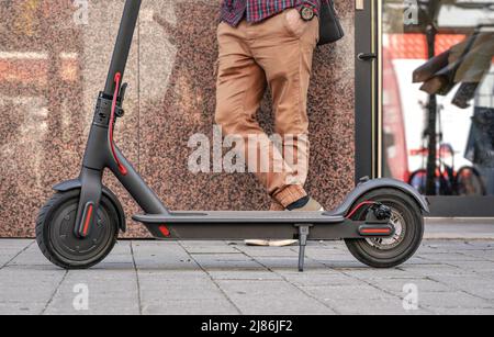 Scooter électrique stationné sur une chaussée carrelée - gros plan, homme debout à côté d'un fond de mur en marbre, seulement ses jambes visibles Banque D'Images