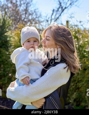 Mère tenant sa fille sur les mains et la regardant avec amour et sourire. Bébé mignon regardant l'appareil photo porter un capuchon et un sweat à capuche. Banque D'Images