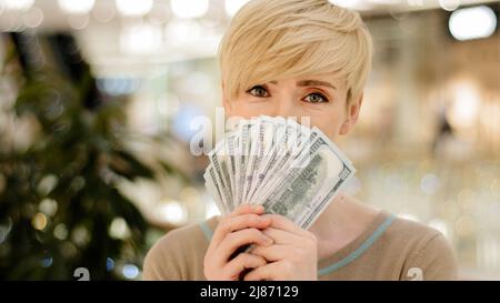Heureux riche adulte d'âge moyen prospère femme d'affaires caucasienne regardant la caméra tenant un fan d'argent se cachant derrière des billets de banque en dollars montrant Banque D'Images