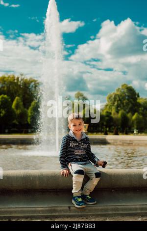 Un petit garçon s'assoit près d'une fontaine par temps ensoleillé. Banque D'Images