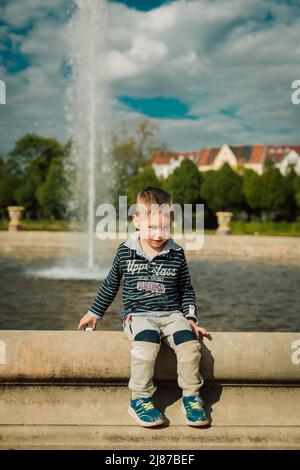 Un petit garçon s'assoit près d'une fontaine par temps ensoleillé. Banque D'Images