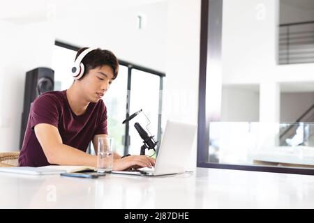 Adolescent asiatique portant un casque avec microphone sur une table et utilisant un ordinateur portable tout en baladodiffusion à la maison Banque D'Images