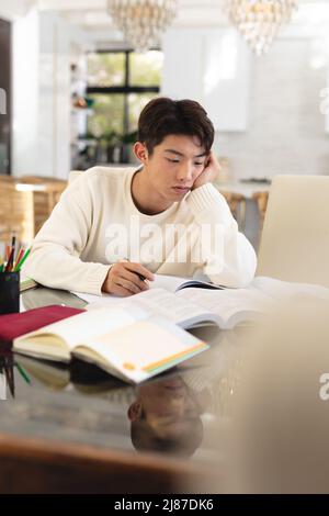 Ennuyé asiatique adolescent garçon avec des livres sur la table étudiant tout en étant assis à la maison, espace de copie Banque D'Images