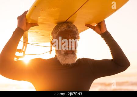Homme senior afro-américain barbu transportant des planches de surf sur la tête à la plage contre un ciel clair au coucher du soleil Banque D'Images