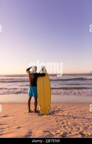 Vue arrière de l'homme d'Amérique africaine avec planche de surf regardant le paysage marin dans un ciel clair Banque D'Images