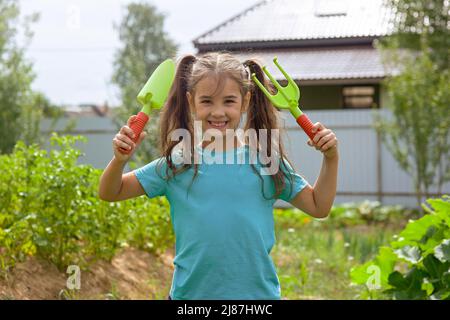 Jolie petite fille tenant de petits outils de jardinage , debout dans le jardin Banque D'Images