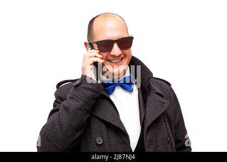 Un homme bald, caucasien adulte habillé avec élégance et portant des lunettes de soleil parle sur son téléphone cellulaire tout en souriant. L'arrière-plan est blanc. Banque D'Images