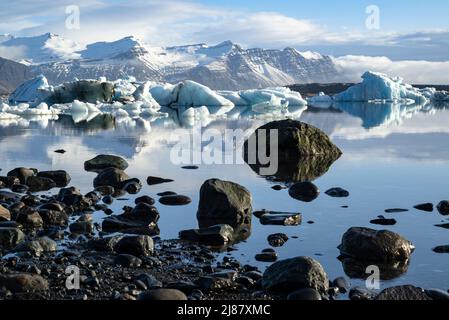 Rive de la lagune de glacier Jökulsárlón avec des rochers noirs et des rochers reflétés dans l'eau et des icebergs flottants au loin, Islande Banque D'Images