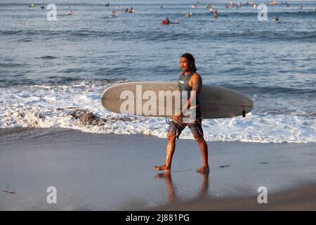 Un surfeur indonésien local marchant le long de la plage de Batu Bolong à Canggu, Bali, Indonésie portant une planche de surf blanche avec des surfeurs dans la mer derrière, Banque D'Images