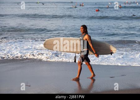 Un surfeur indonésien local marchant le long de la plage de Batu Bolong à Canggu, Bali, Indonésie portant une planche de surf blanche avec des surfeurs dans la mer derrière, Banque D'Images