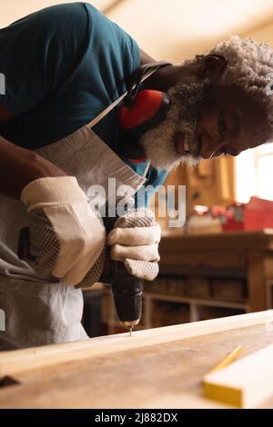 Souriant menuisier afro-américain mature faire le trou avec le foret sur la planche à l'établi dans l'atelier Banque D'Images