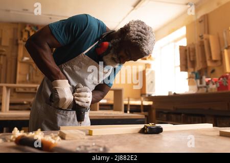 Menuisier afro-américain mature faisant le trou avec le foret sur la planche à l'établi dans l'atelier Banque D'Images