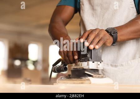 Section médiane du menuisier africain américain mature utilisant une ponceuse sur la planche à l'atelier Banque D'Images