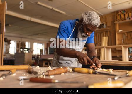 Charpentier africain américain mature utilisant le burin sur la planche dans l'atelier de travail du bois Banque D'Images