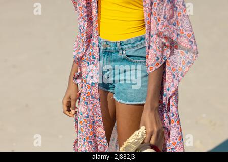 Section médiane de la jeune femme afro-américaine portant une veste sarong et un pantalon chaud tenant un chapeau à la plage Banque D'Images