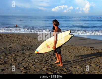 Un surfeur indonésien local marchant le long de la plage de Batu Bolong à Canggu, Bali, Indonésie portant une planche de surf blanche avec des surfeurs dans la mer derrière, Banque D'Images