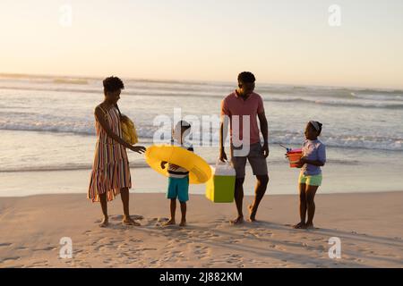 Parents afro-américains regardant son fils et sa fille jouer avec anneau gonflable et seau sur la plage Banque D'Images