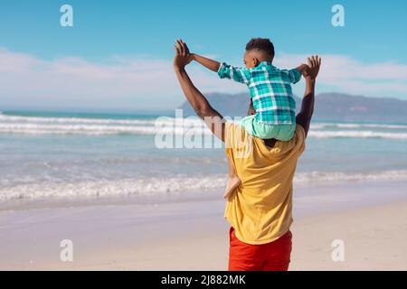 Vue arrière du jeune homme afro-américain avec les bras levés portant son fils sur les épaules à la plage Banque D'Images