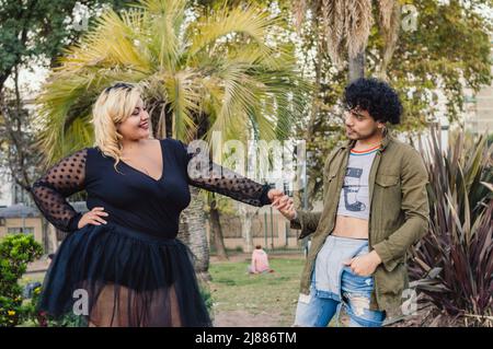 deux jeunes amis caucasiens, une femme de plus grande taille et un homme avec des curlers, tenant les mains face à l'autre, posant à l'extérieur du parc, avec des arbres dans le Banque D'Images