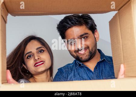 Vue en coupe basse de l'intérieur de la boîte gros plan: Happy indian couple ouvrant la boîte en carton après le déplacement et regardant à l'intérieur dans la nouvelle maison Banque D'Images