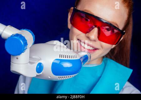 gingembre rougeâté jeune cliente gaie avec des lunettes rouges de protection assises dans le cabinet dentaire et obtenir un sourire blanchissant dans la chaise d'oeuf Banque D'Images