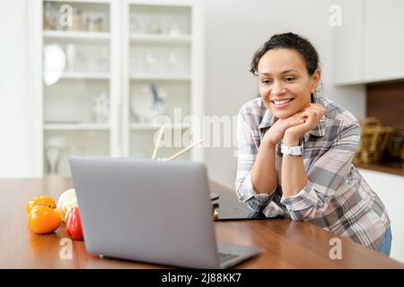 Bonne belle jeune femme appréciant la cuisine, à la recherche d'une recette sur l'ordinateur portable dans la cuisine ordinateur portable sur la table de cuisine et la cuisine. Concept de blogueur de nourriture Banque D'Images