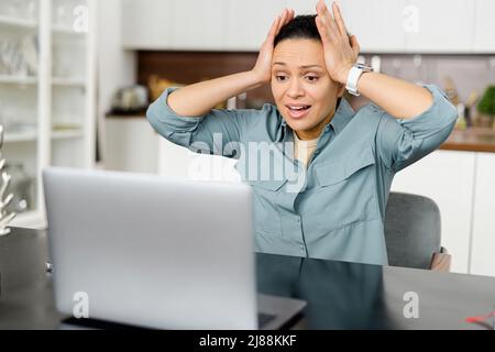 Une femme frustrée se trouve devant l'ordinateur portable et regardant l'écran avec une expression de visage choquée, mécontent de mauvaises nouvelles, la femme tient la tête avec les deux mains, a un problème avec le travail Banque D'Images