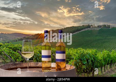 Un gobelet en verre avec vin blanc et deux bouteilles sur un baril, avec le fond du vignoble au coucher du soleil, Toscane, Italie Banque D'Images