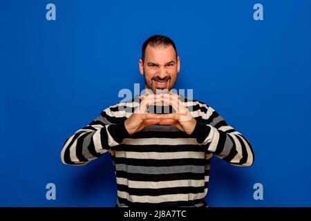 Latino homme avec la barbe mettant ses mains ensemble diabolement montrant ses mauvaises intentions, isolé sur fond bleu studio Banque D'Images