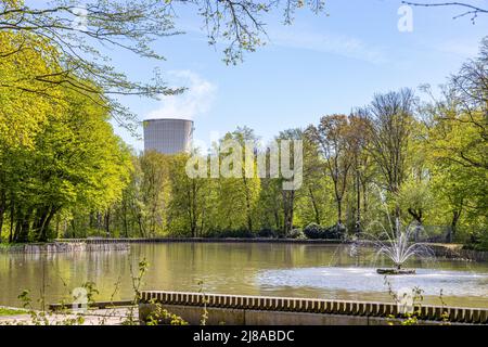 Étang avec une petite fontaine, arbres verts luxuriants et une énorme cheminée d'un complexe industriel en arrière-plan, jour ensoleillé avec un ciel bleu de source à Heidek Banque D'Images