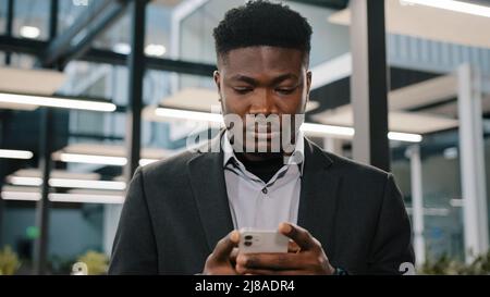 Un jeune homme sérieux travailleur de bureau afro-américain à la recherche d'un smartphone Internet en choisissant de passer commande en discutant en ligne avec des amis en défilement mobile Banque D'Images