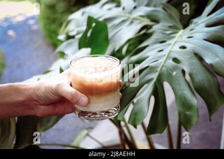 Tenez le café latte glacé à la main dans un grand verre. Boisson d'été froide avec fond vert nature Banque D'Images