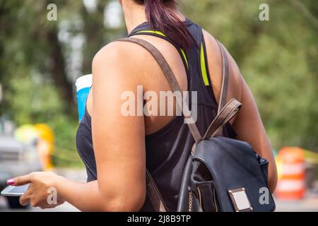 Une femme écourtée et méconnaissable avec un t-shirt d'été et un sac à dos en cuir marron et noir tient une boisson sans alcool et un téléphone portable sur fond de bokeh Banque D'Images