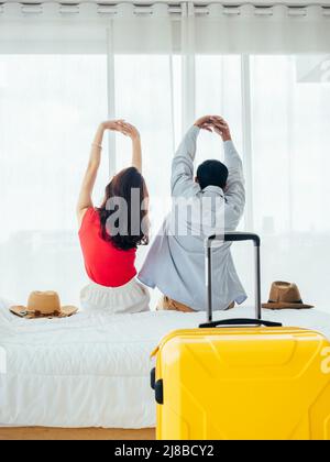 Couple de touristes, de joyeuses vacances. Vacances d'été. Vue arrière de jeune asiatique homme et femme s'étirant avec détente sur le lit avec valise près du rideau Banque D'Images