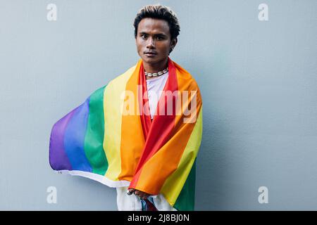 Jeune homme enveloppé dans le drapeau LGBT arc-en-ciel debout sur le mur gris à l'extérieur Banque D'Images