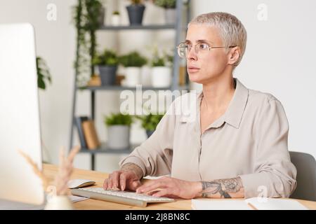 Femme d'affaires d'âge moyen concentrée dans des lunettes de vue assis au bureau et tapant sur le clavier tout en correspondant avec le partenaire d'affaires Banque D'Images