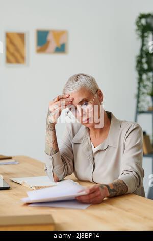Femme d'affaires d'âge moyen attentionnés aux cheveux blonds assise à table et examinant la lettre d'affaires Banque D'Images