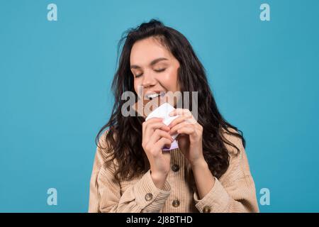 La jeune femme européenne brune heureuse avec les yeux fermés mange du chocolat et apprécie le goût des bonbons Banque D'Images