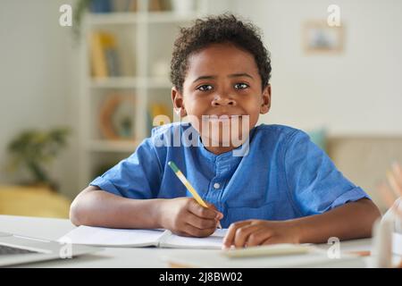 Portrait d'un garçon afro-américain souriant en chemise bleue, assis à table et prenant des notes dans un cahier d'exercices Banque D'Images