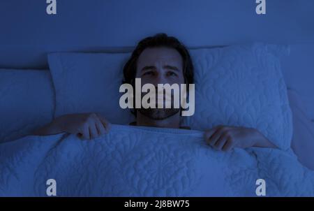 Un jeune homme européen déprimé avec un chaume se trouve sur un lit blanc sous une couverture dans la chambre à l'intérieur à l'obscurité Banque D'Images