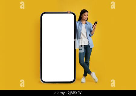 Bonne jeune japonaise femme dans un look décontracté au smartphone, se tient près d'un énorme téléphone avec écran vide Banque D'Images