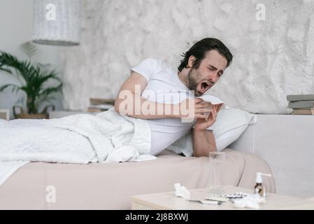 Triste millennal caucasien gars avec chaume se trouve sur le lit, éternue à la serviette dans la chambre à l'intérieur avec des médicaments Banque D'Images