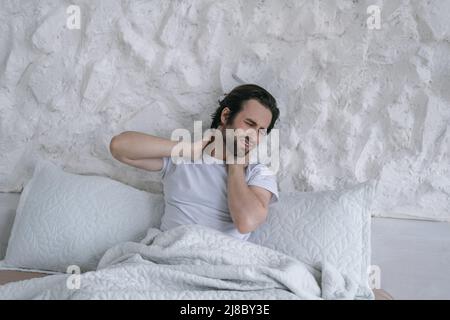 Triste triste millennium gars caucasien avec le chaume se réveiller, assis sur le lit et souffrant de douleur dans les muscles de cou Banque D'Images