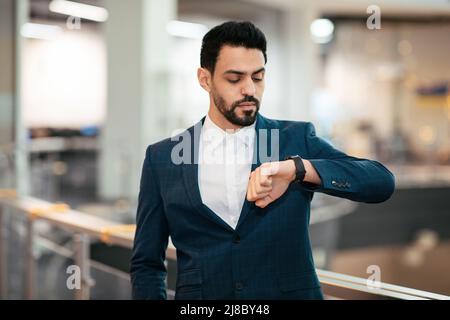 Sérieux calme attrayant jeune homme de pdg arabe avec la barbe en costume regarde la montre intelligente attendre le client à l'intérieur du bureau Banque D'Images