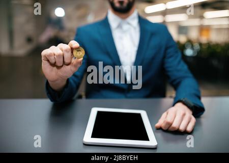 Homme en costume avec montre intelligente tenir le bitcoin en main s'assoit à la table avec une tablette dans l'intérieur du bureau Banque D'Images