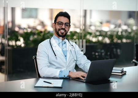 Souriant beau jeune homme arabe médecin avec une barbe en lunettes et un manteau blanc, au travail travail avec un ordinateur portable Banque D'Images
