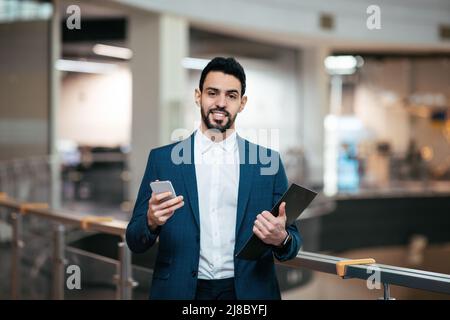 Joyeux homme du Moyen-Orient millénaire avec barbe en costume utilisez une tablette et un smartphone à l'intérieur du bureau Banque D'Images