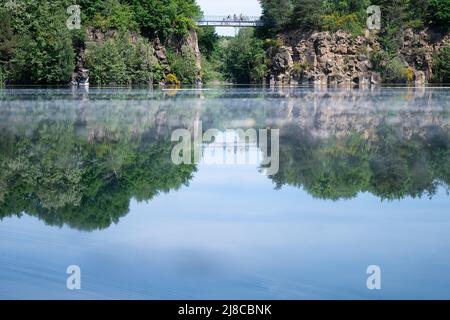 15 mai 2022, Hessen, Mühlheim am main: Le ciel bleu et les murs de pierre d'une ancienne carrière, entre laquelle un pont est étiré, se reflètent dans le lac Vogelsberg. En 1982, un paysage lacustre d'une superficie totale de plus de 61 hectares a été créé dans les anciennes carrières de basalte. Photo: Sebastian Gollnow/dpa Banque D'Images