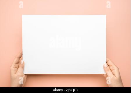 Les mains des femmes tiennent une feuille de papier vierge sur un fond beige. Les mains des femmes avec une feuille de papier vierge sur une table rose. Mains humaines tenant la maquette blanc adv Banque D'Images